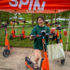A woman stands under a red SPIN-branded canopy, smiling while holding an orange SPIN electric scooter. She's wearing glasses, a green sweatshirt, shorts, and sandals, and has a beige tote bag on her arm. Behind her, several more scooters are lined up on g